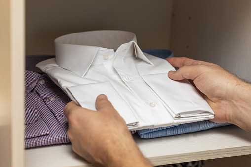 A man puts a new folded long sleeve white shirt in an open shelf in his closet