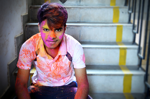 Horizontal portrait of one happy smiling young 16 year old boy with smile on his face painted messy with Holi powder colours sitting on gray colored rustic stairs in the background