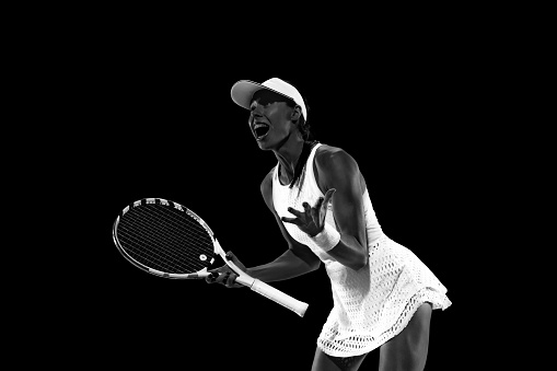 Successful match. Tennis player holds racquet and expressing joy and happiness against black studio background. Monochrome filter. Concept of women in sport, active lifestyles, energy, movement.