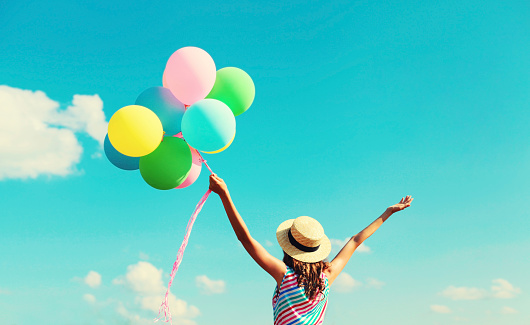 Back view of happy joyful young woman with bunch of colorful balloons in summer straw hat on blue sky clouds background