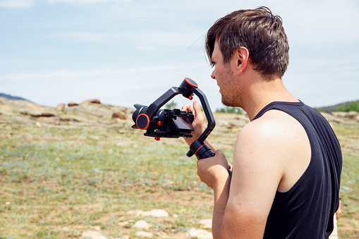 Young adult man shooting video on camera with handheld electronic stabilizer on nature in mountains in summer. portrait of man videographer while working