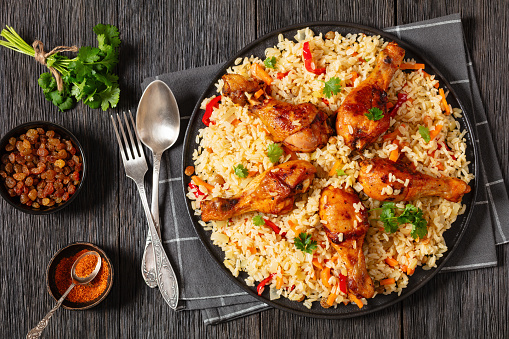 fried chicken drumsticks over brown rice pilaf with vegetables and raisins on black platter on dark wooden table with spoon, fork and ingredients, horizontal view from above, flat lay, free space