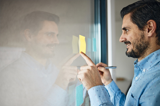 Photo of a young man writing on a transparent wipe board and thinking of a solution for his work-related problems.