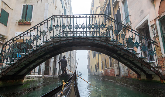 This is an image of a traditional gondola being punted by a traditional gondolier going under one of the many canal bridges in Venice