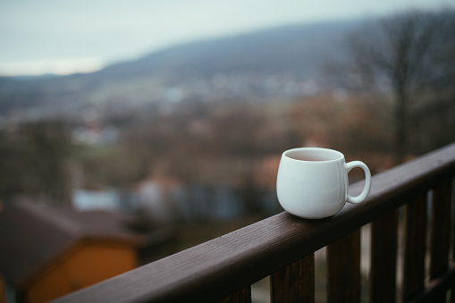 White cup with hot fresh tea on wooden balcony with mountains view.