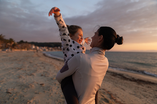 Mother carrying and hugging her little daughter by the sea at sunset