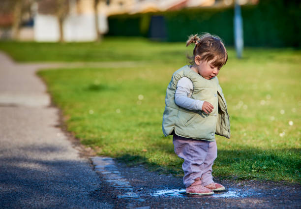 Rascal little female child with braids jumping in a muddy puddle on a sunny spring day. Scoundrel 2 year old girl hopping in a small water plash by the grass. Kids exploration and freedom concept - Malmo, Sweden