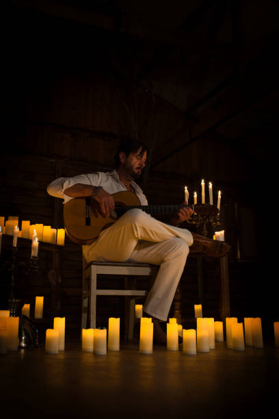 istock Man dressed in white enjoying playing Spanish guitar at home by candlelight, learning musical instrument, musician or singer singing new single song. Vertical photography. 2039844081
