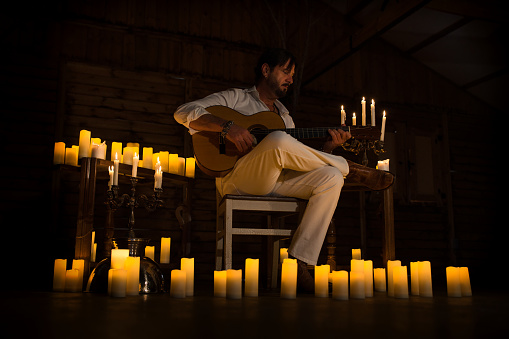 Man dressed in white enjoying playing Spanish guitar at home by candlelight, learning musical instrument, musician or singer singing new single song.