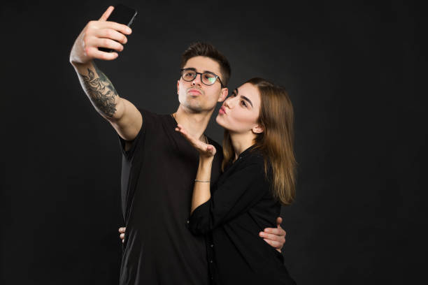 happy young loving couple making selfie and smiling while standing against black background. - adult isolated color image studio shot stock-fotos und bilder