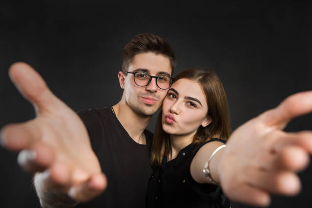 happy young loving couple making selfie and smiling while standing against black background. - adult isolated color image studio shot stock-fotos und bilder
