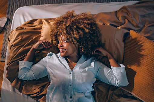 High angle view of a happy woman with curly hair, smiling and lying in a cozy bed with stylish pillows and bedding.
