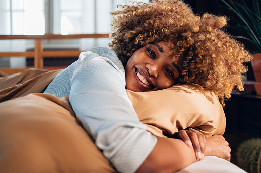 Smiling African American woman with curly hair hugging a cushion on a comfortable sofa, feeling content and relaxed at home.