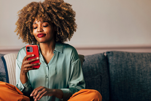 A stylish young woman with curly hair browses her phone with a contented expression while sitting on a sofa.