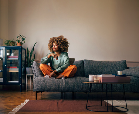 Fashionable young woman with curly hair sitting pensively on a couch, surrounded by indoor plants and contemporary decor.