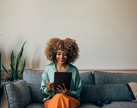 Cheerful young woman with curly hair enjoying digital content on a tablet while sitting comfortably on a couch.