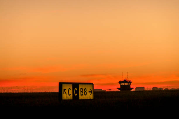 A taxiway direction sign for pilots at Sydney Kingsford-Smith Airport. In the background is the old air traffic control tower at Kyeemagh Beach.  The silhouette of spikes to deter birds are visible along the top of the sign. In the far distance are residential buildings.  This image was taken from near Shep's Mound, Ross Smith Avenue, Mascot, on a hot, sunny day after sunset on 25 February 2024