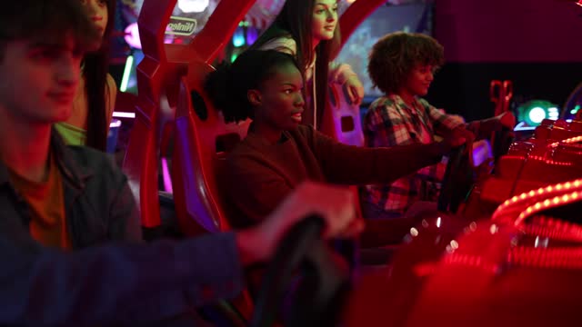 group of multiracial teenage friends in arcade playing car games with steering wheel