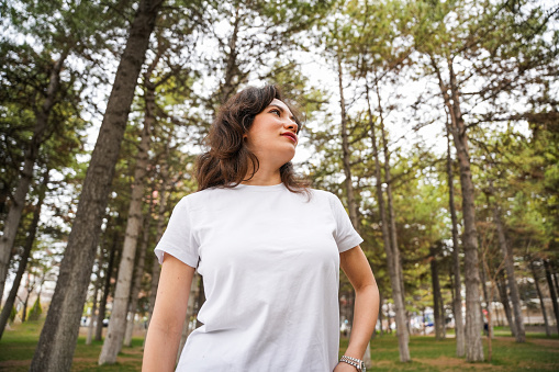 The woman in a white T-shirt is standing in front of the pine trees, gazing at the forest
