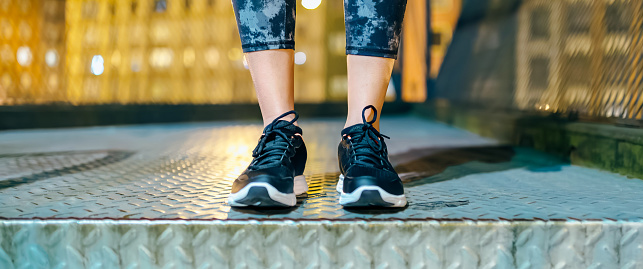 Banner of unrecognizable female athlete legs wearing leggings and sneakers ready to night run on streets. Sportive woman feet standing on metallic urban stair with illuminated city on background.