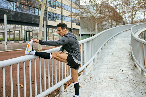 Side view of man in late 40s wearing sports clothing, using footbridge railing to warm up before running.