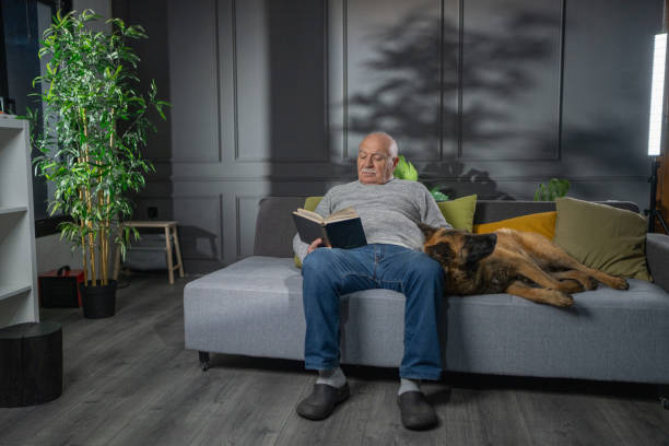 Senior man relaxing while sitting on sofa, reading a book and petting his dog in the evening at home