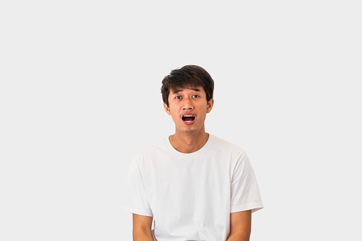 A young Asian man in his 20s wearing a white t-shirt is looking shocked at what is in front of him, isolated on a gray background.