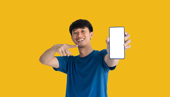 A young Asian man in his 20s wearing a blue t-shirt smiling confidently showing mobile phone with blank white screen to camera, standing isolated on yellow background. Closed smartphone in man's hand