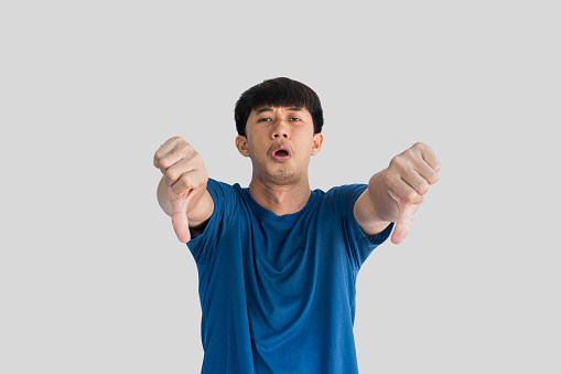 A young Asian man in his 20s wearing a blue t-shirt making a displeased gesture showing a thumbs down isolated on a gray background. The concept of expressing dislike or opposing opinions.