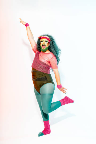 Full body of positive young beaded crossdresser man in colorful body fit female leggings and top with eyeglasses looking at camera while dancing with twisted hands against white background
