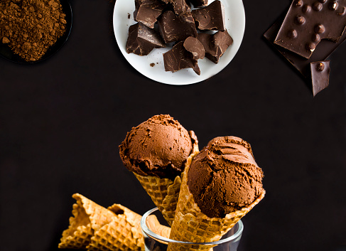 Chocolate ice cream in a waffle cone, piece of dark chocolate and cocoa powder on the black background. Close-up.