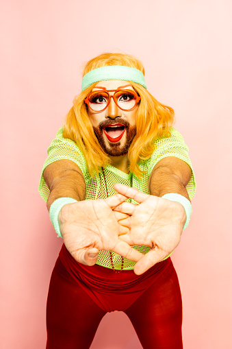 A drag queen clad in vibrant 80s exercise attire, including a neon headband, performs a stretch with a playful expression