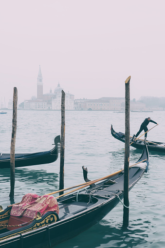 Venice, Italy - February 19, 2022: A Venetian gondolier steers his craft past other gondolas which are tied up on the side of a misty Grand Canal in Venice. Across the water we see the iconic shape of the Church of San Giorgio Maggiore