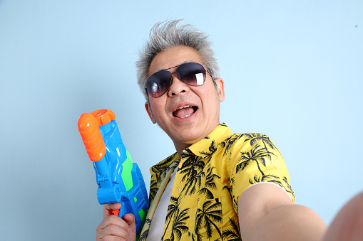 HAPPY SONGKRAN DAY. Asian energetic senior man in summer clothing and sunglasses with gesture of holding plastic water gun while take a selfie isolated on blue background. Songkran festival. Thai New Year's Day.