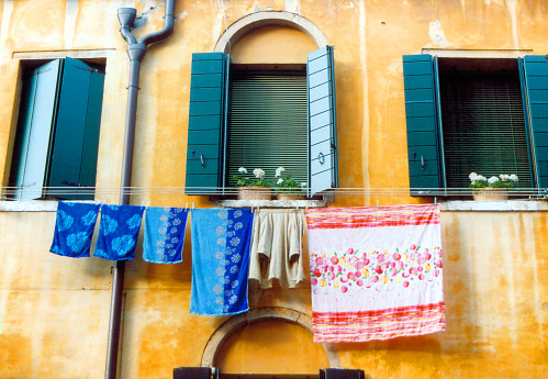 Venice, Veneto, Italy: façade with laundry - old yet charming building with a warm yellow façade. Three green window shutters are slightly ajar, below, clothes hang to dry, painting a picture of ordinary moments made extraordinary by the dance of colors and textures.