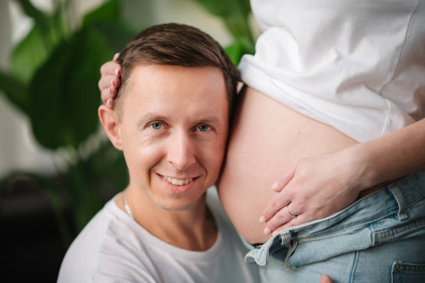 남편이 임신한 아내의 뱃속에서 아기를 듣고 있는 모습. 임산부와 사랑스러운 잘생긴 남자가 집에서 배를 껴안고 있습니다. 부모의 개념. 아기를 기다리는 커플. 9개월 클로즈업. - human pregnancy baby shower image color image 뉴스 사진 이미지