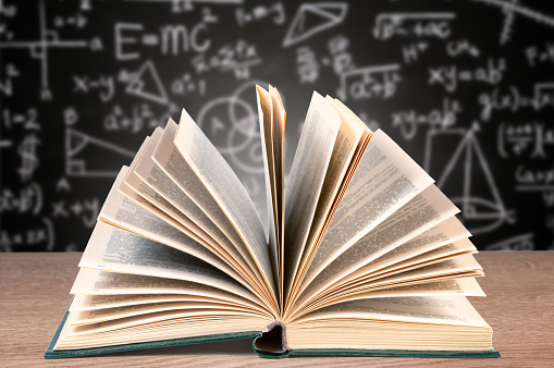 Open Textbook on Desk Against Blurred Blackboard With Mathematical Equations