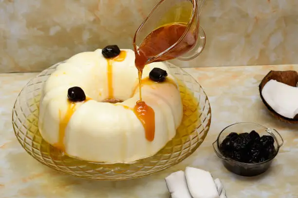Manjar decorated with plums, the syrup is poured into a slice on a white plate, on a yellow marble background