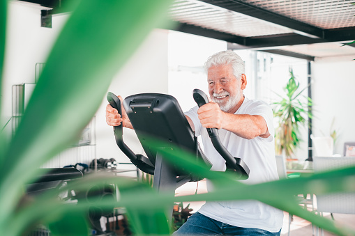 People sport concept. Happy senior man cycling doing exercises to stay fit. Elderly bearded man riding stationary bicycle in home terrace