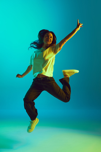 Full-length portrait of young beautiful woman jumping raising hands of joy in neon yellow lighting against gradient blue studio background. Concept of emotions, facial expression, self-expression. Ad