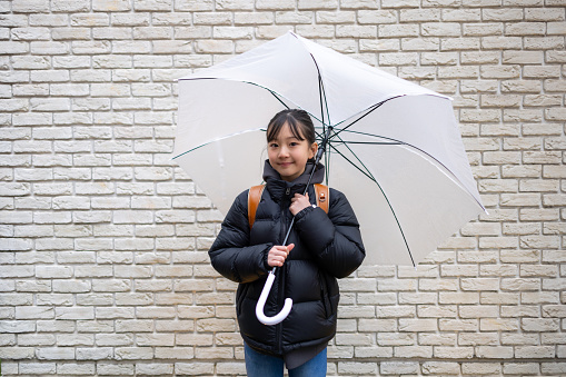 Portrait of Japanese elementary school student girl carrying a ‘randoseru’ school bag in a rainy day, standing against white wall