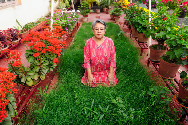 Full-body view of woman in her 60s wearing white T-shirt and black pants, walking on green lawn among blooming plants. Scene of peaceful outdoor life and slow-paced recovery.