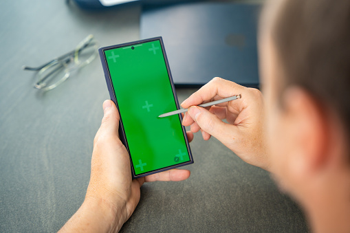 Close up view of man hand holding smart phone with green chroma key screen and stylus pen at his creative workspace. High quality photo