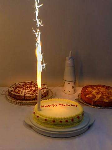 Birthday cake buffet with traditional cakes. One cake with yellow frosting and red decoration has the handwritten words „Happy Birthday“ on it.  A real fireworks candle burns and sprays glittering sparks.