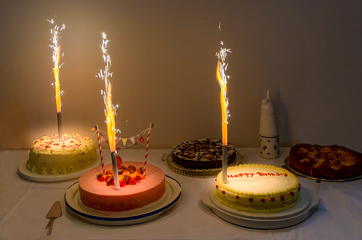 Birthday cake buffet with traditional homemade cakes. A cake with red icing is beautifully decorated. One cake with yellow frosting and red decoration has the handwritten words „Happy Birthday“ on it. Real fireworks candles burn and spray glittering sparks.