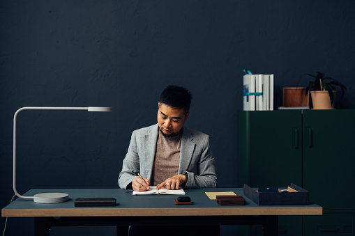 A businessman in a suit is focused on writing notes at a minimalist workspace with soft lighting.