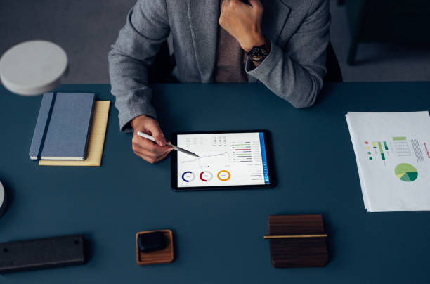 Professional Analyzing Financial Reports with Tablet at Office Desk Focused professional at work analyzing financial reports and graphs on a tablet, with notebooks and papers on desk. planned economy stock pictures, royalty-free photos & images