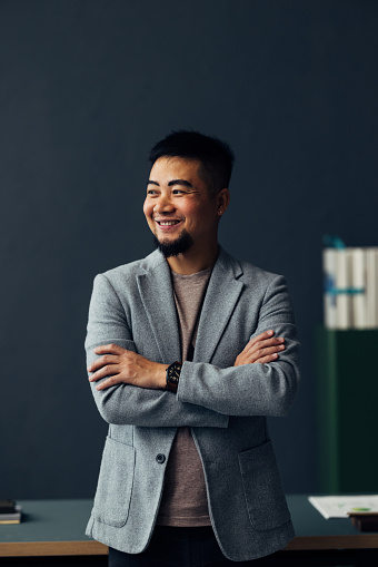 Portrait of a cheerful Asian man wearing business casual attire, standing in a modern office environment with a genuine smile.