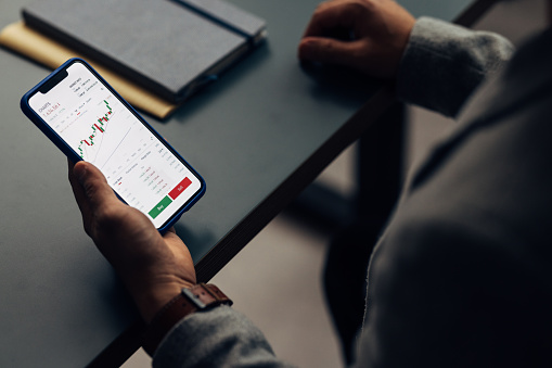 Close-up of a professional's hand holding a smartphone displaying stock market charts, with a notebook in the background.