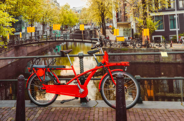 A close-up view captures the quintessential scene of Amsterdam, with a parked bicycle in the foreground and the picturesque backdrop of canals, trees, cars, and historical buildings. The bicycle, a symbol of the city's cycling culture, stands prominently against the charming streetscape, while the tranquil canal and lush greenery provide a serene atmosphere. In the distance, the architectural marvels of Amsterdam's traditional buildings add to the timeless beauty of this iconic European cityscape.
